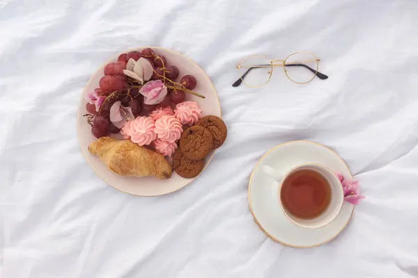 stock image A romantic flat lay features a cup of tea, a plate with cookies, a croissant, pink meringues, and grapes on a white blanket. A pair of glasses adds a personal touch, creating an elegant and serene