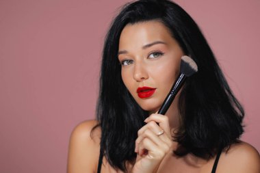 Kırmızı dudaklı ve siyah saçlı çekici bir kadın pembe arka planda yanağına makyaj fırçası tutuyor. Makyaj dersleri, güzellik terfileri ve kozmetik aletleri reklamları için ideal.