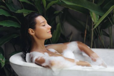Canlı tropikal bitkilerle çevrili, açık havada lüks bir küvette banyo yapan bir kadının huzur dolu bir sahnesi. Bu görüntü bir rahatlama ve kendine bakım hissi uyandırıyor, spa ve sağlık için ideal.
