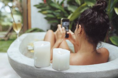 Açık havada banyoda dinlenen, etrafı mumlarla çevrili akıllı telefonunu kullanan bir kadının huzurlu görüntüsü. Bu fotoğraf modern teknolojinin ve barışçıl lüksün bir karışımını yakalıyor.
