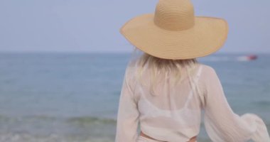 Güneş şapkalı gülümseyen bir kadın sahilde poz verir ve döner, yaz neşesi ve modasının özünü yakalar. Seyahat, rahatlama ve deniz kenarı zarafeti temaları için mükemmel..