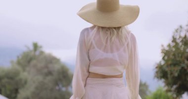 Geniş kenarlı güneş şapkalı şık sarışın kadın manzaralı bir ortamda zarafetle dönüyor. Moda, yaşam tarzı ve seyahat içeriği için mükemmel. Yüksek kaliteli görüntüler yaz zarafetini yakalıyor..