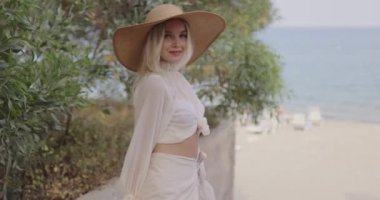 Güneş şapkalı sarışın bir kadın sahilde geziniyor, deniz kenarında sakin ve şık bir anı yakalıyor. Yaz modası, seyahat ve rahatlama temaları için ideal.