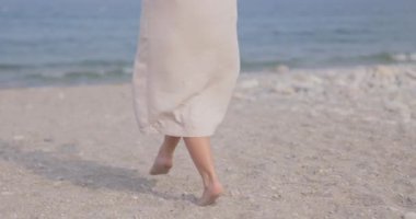 Saf kıyafetli bir kadın bir çakıl taşı plajı boyunca yürüyor, sakin sahil ortamının tadını çıkarıyor. Moda, rahatlama ve sakin sahil yürüyüşleri temaları için mükemmel..