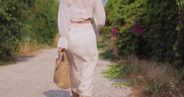 Şık bir yaz elbisesi içinde zarif bir kadın ve büyük bir güneş şapkası ile pitoresk bir patikada yürüyor. Yaşam tarzı, moda ve seyahat içeriği için ideal. Yüksek kalite video gösterimi yaz zarafeti.