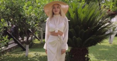 Geniş kenarlı güneş şapkalı şık sarışın kadın yemyeşil bir bahçede zarafetle yürüyor. Moda, yaşam tarzı ve seyahat temaları için ideal. Yüksek kaliteli görüntüler yaz zarafetini yakalıyor..