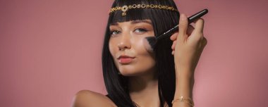 Kleopatra 'dan esinlenilmiş altın başlıklı ve parlak siyah saçlı güzel bir kadın pembe arka planda makyaj fırçasıyla pudra sürüyor. Özgüvenli ifadesi ve lüks stili şu temaları temsil ediyor: