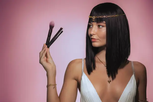 stock image A stylish woman with sleek black hair, adorned with a Cleopatra-inspired gold headpiece, holds up makeup brushes against a pink background. Her poised expression and luxurious appearance make this