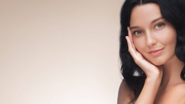 Koyu saçlı, kendine güveni ve doğal güzelliği olan zarif bir kadının stüdyo portresi. Yumuşak bir bej arka plan karşısında eli yüzü üzerinde zarifçe poz veriyor. Benlik teması için mükemmel