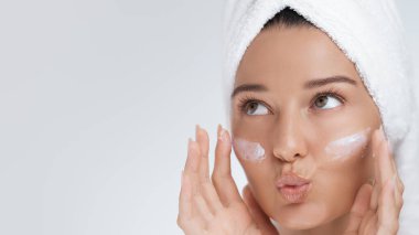 Yüzüne havluyla sarılmış gülümseyen bir kadın yüzüne cilt kremi sürerken neşeli bir yüz ifadesi takınıyor. Güzellik, özgüven ve neşe temalarını resmetmek için mükemmel.