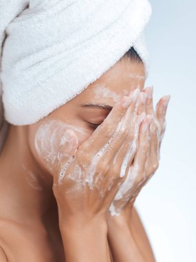 Havluya sarılmış bir kadın sabah rutininin bir parçası olarak yüzünü köpüklü temizleyiciyle kaplıyor. Bu görüntü güzellik, hijyen, sağIık ve kişisel bakım kavramlarını desteklemek için mükemmeldir..