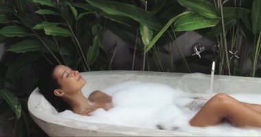 Huzurlu bir kadın, bereketli tropikal bitkilerle çevrili lüks bir köpük banyosunda uzanır. Bu barışçıl görüntü sağlık, spa ya da rahatlama temalı projeler ve promosyonlar için mükemmeldir..