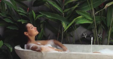 Lüks bir köpük banyosunda uzanan bir kadın, açık hava spa ortamında yemyeşil tropikal bitkilerle çevrili. Sağlık, güzellik ve tatil kampanyaları için ideal. Yüksek kaliteli yavaş çekim görüntüleri mükemmel.