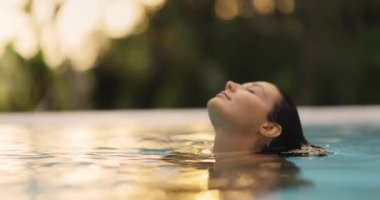 Tropik cennet, gün batımında göğsünü elleriyle kaplayan sonsuz havuzda seksi turuncu mayo giymiş, bikinili esmer kadının lüks rahatlığı. Yüksek kalite ağır çekim 4K görüntü