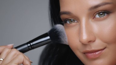 Kusursuz cildini geliştirmek için yumuşak bir makyaj fırçası kullanan ışıl ışıl bir kadının yakın çekimi. Güzellik reklamları, kozmetik dersleri ve cilt bakımı için mükemmel..