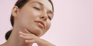 Kapalı gözlü bir kadının çenesine hafifçe dokunduğu, rahatladığı ve kendine baktığı bir anın tadını çıkardığı yakın plan. Yumuşak pembe arka plan dingin ve rahatlatıcı atmosferi arttırır..