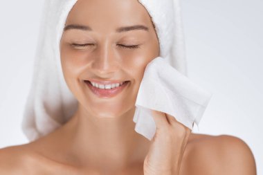 Kafasına havlu sarılmış gülümseyen bir kadın yüzünü bir yüz silme ile nazikçe temizliyor, ferahlatıcı bir cilt temizleme rutininin keyfini çıkarıyor. Güzelliği, kendine bakımı, hijyeni ve yüz bakımını teşvik etmek için ideal.