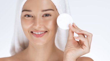 Kafasına havlu sarılmış mutlu bir kadın elinde pamuk tutarak cilt bakımının keyfini çıkarıyor. Güzellik, hijyen, kendine bakım ve yüz bakımı kavramlarını göstermek için mükemmel..