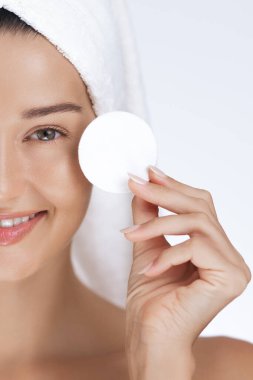 Saçına havlu sarılmış gülümseyen bir kadının yakın plan fotoğrafları. Yüzüne pamuk tutuyordu. Cilt bakımına hazırdı. Güzellik, kendine bakım ve yüz bakımı kavramları için ideal.