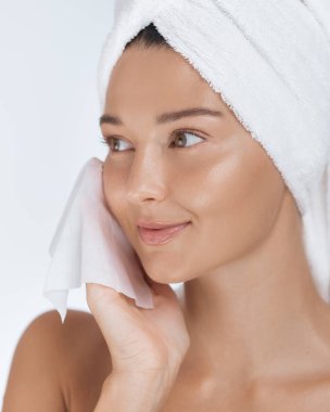 Havluya sarılmış bir kadın yüzünü temizlemek için yumuşak bir bez kullanır. Parlak cildini ön plana çıkarır. Güzellik, cilt bakımı ve cilt bakımını teşvik etmek için ideal, taze cilt ve ışıl ışıl