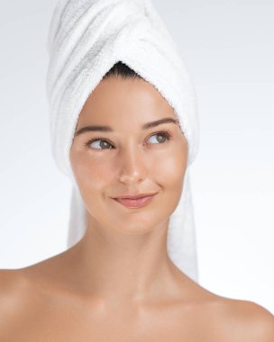 Taze, parlak bir teni olan bir kadın beyaz bir havluya sarılı iken düşünceli bir şekilde gülümsüyor. Cilt bakımını, güzelliği, refahı ve kendine bakımı teşvik etmek için idealdir.