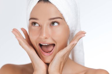 Kafasına havlu sarılmış neşeli genç bir kadın ellerini yüzünü çerçeveletip sürpriz ve heyecan sergiliyor. Cilt bakımını, güzelliği, sağlığı ve kişisel bakım ürünlerini teşvik etmek için mükemmel.
