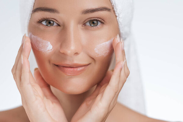 Крупный план улыбающейся женщины с сияющей кожей, наносящей увлажняющий крем на щеки, подчеркивающий повседневную рутину кожи. Идеально подходит для продвижения красоты, лыж и велнес.