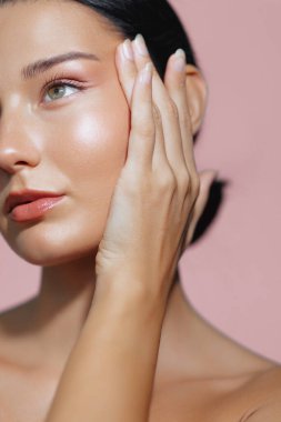 Stunning Latina woman with radiant skin and natural makeup poses with her hand gently touching her face in this close-up portrait against a pink background. Ideal for skincare promotions, beauty clipart