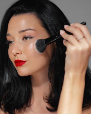 Kalın kırmızı dudaklı zarif bir kadının yakın çekimi. Fırçayla makyaj yapıyor. Kusursuz bir ten ve ışıl ışıl bir güzelliğe vurgu yapıyor. Kozmetik, cilt bakımı ve moda reklamları için ideal.