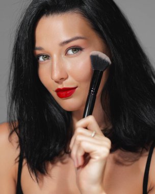 Kendine güvenen kırmızı dudaklı, yüzünde makyaj fırçası tutan, kusursuz bir ten ve güzellik gösteren bir kadının yakın çekimi. Kozmetik, makyaj dersleri ve cilt bakımı reklamları için mükemmel.