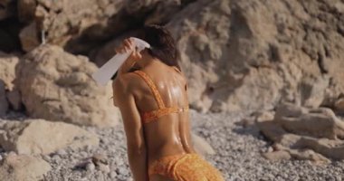 Bikinili fit bir kadın kayalık bir sahilde güneşlenirken su spreyiyle serinliyor. Yaz, seyahat ve yaşam tarzı içeriği için ideal. Yüksek kaliteli stok görüntüleri.