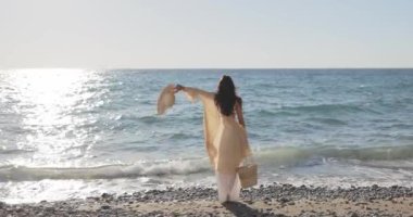 Gün batımında dalgalı bir örtüyle okyanusa doğru yürüyen bir kadının arka görüntüsü, bir elinde şapkasıyla. Huzur, özgürlük, yaz tatili ve deniz kenarı kaçışları temaları için mükemmel..