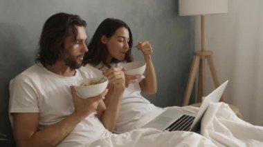 Bir çift laptopta bir şey izlerken yatakta kahvaltıdan zevk alır, rahat bir sabah rutini yakalarlar. Sinema yavaş çekim 4k video, reklam için tasarım ve sosyal medya, vlog