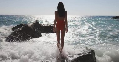 Kırmızı mayo giymiş bir kadın, etrafındaki okyanus dalgalarıyla kıyı kayalıklarında güvenle duruyor. Bu dramatik sahne, gücün, maceranın ve denizin saf güzelliğini yakalar..