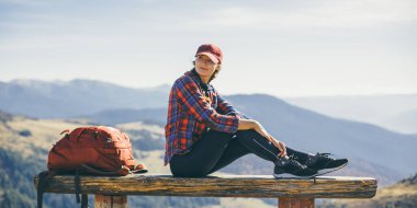 Kırmızı sırt çantalı bir bayan yürüyüşçü bir bankta mola verir, manzaralı dağ manzarasının tadını çıkarır. Bu yüksek kaliteli görüntü seyahat, macera ve doğa keşfi kavramları için mükemmeldir..