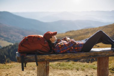 Kırmızı sırt çantalı bir bayan yürüyüşçü, dağ manzarasının nefes kesen manzarasının tadını çıkarırken bir bankta uzanır. Bu yüksek kaliteli görüntü seyahat, rahatlama ve rahatlama kavramları için idealdir.
