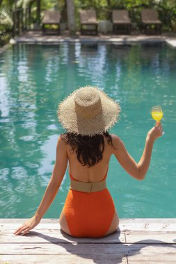 Turuncu bikinili bir kadın, elinde ferahlatıcı bir içecek tutan ve büyük bir şapka takan bir havuzun kenarında oturur. Sakin tropikal ortam rahatlama ve rahatlama için mükemmel bir zemin sağlar..