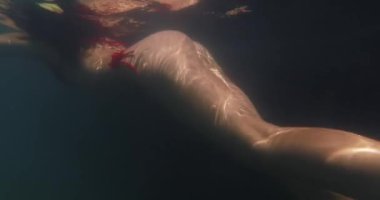 Derin okyanusta tenine yansıyan güneş ışığıyla kırmızı bikinili bir kadının şehvetli sualtı görüntüsü. Lüks, egzotik seyahat ve deniz kenarında yaz tatili kavramları için idealdir..