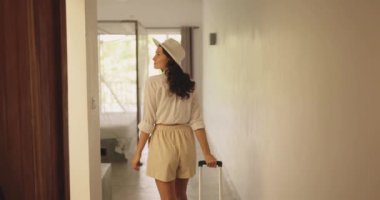 Yaz kıyafeti giymiş, şapkalı bir kadın parlak bir otel odasına elinde bavulla giriyor. Seyahat, tatil ve konukseverlik için mükemmel promosyonlar veya kavramlar.