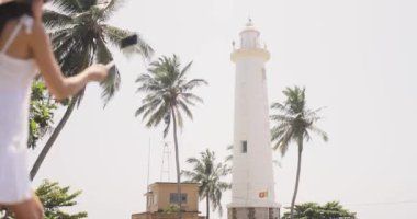 Beyaz elbiseli ve güneş şapkalı bir kadın, Sri Lanka 'daki Galle Fort deniz fenerinin yakınlarında vloglara biniyor. Bu yüksek kaliteli görüntüler, turizm için mükemmel olan seyahat ve sosyal medya içeriği oluşturma karışımını yakalıyor.