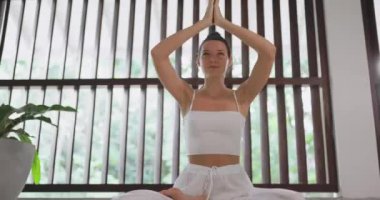 Huzurlu bir kadın, meditasyon pozisyonunda bağdaş kurarak oturur. Huzurlu bir yerde, beyaz yoga kıyafeti giyerek. Farkındalık, sağlık, meditasyon ve yoga uygulamalarıyla ilgili içerik için ideal.