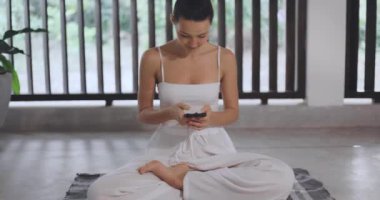 Gülümseyen bir kadın yoga pozunda oturuyor, elinde yeşil ekranlı bir akıllı telefon ve huzurlu bir ev ortamında. Mobil uygulamaları, meditasyon araçlarını veya refahla ilgili teknolojiyi göstermek için mükemmel..