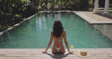 Beyaz bikinili bir kadın lüks bir tropikal havuzun kenarında oturuyor. Rahatlatıcı tatil sahnesi egzotik yerlerde seyahat, boş zaman, lüks veya yaz kaçamakları için mükemmeldir..