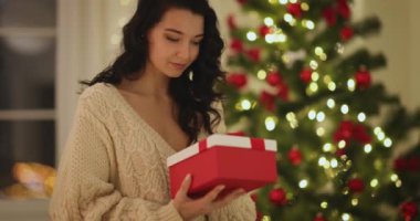 Uzun siyah saçlı genç bir kadın güzel dekore edilmiş bir Noel ağacının kırmızı hediye kutusunu neşeyle açıyor. Bu festival görüntüsü, tatil sezonunun sıcaklığını ve heyecanını yansıtıyor.