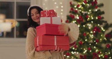 Güzel ışıklandırılmış ve dekore edilmiş bir ağacın önünde Noel Baba şapkalı neşeli bir kadın bir yığın hediyeyi kucaklıyor. Tatil promosyonları ve şenlikli pazarlama için ideal..