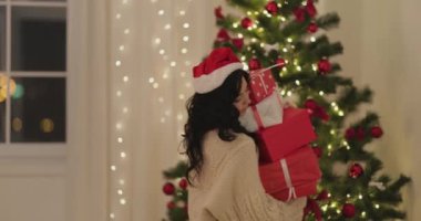 Noel Baba şapkası takan ve rahat süveter giyen bir kadın güzel dekore edilmiş ışıklarla süslenmiş bir ağacın yanına Noel hediyelerini dikkatlice yerleştiriyor. Tatil promosyonları ve festival reklamları için mükemmel..