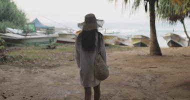 Plaj elbiseli ve güneş şapkalı bir kadın sakin bir Sri Lanka plajında teknelere doğru yürüyor. Seyahat promosyonlarında, tropikal tatil reklamlarında ve yaşam tarzı içeriğinde kullanım için idealdir. Huzurlu sabah sahnelerini yakalar.. 