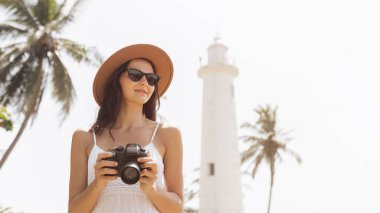 A female tourist in sunglasses and a hat holds a camera while standing in front of the Galle Fort lighthouse in Sri Lanka. The tropical background with palm trees and clear skies evokes travel and clipart