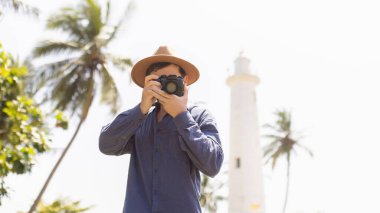 Geniş kenarlı şapkalı bir erkek fotoğrafçı Sri Lanka 'daki Galle Fort deniz fenerinin yakınında bir resim çekiyor. Palmiye ağaçları ve açık gökyüzü ile çevrili tropikal ortam seyahat için idealdir.