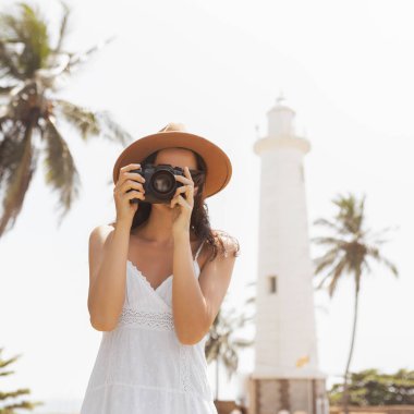Şapka ve beyaz elbise giyen bir kadın fotoğrafçı Galle Fort, Sri Lanka 'daki deniz fenerinin fotoğrafını çekiyor. Palmiye ağaçları ve güneş ışığıyla tropikal arka plan seyahat ve macerayı geliştirir.